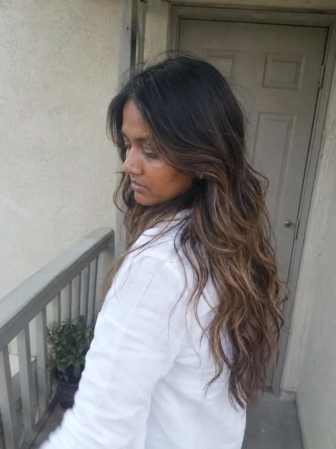A bright indoor selfie of a smiling woman looking directly into the camera, showcasing a textured, shoulder-length wavy lob haircut with caramel blonde highlights