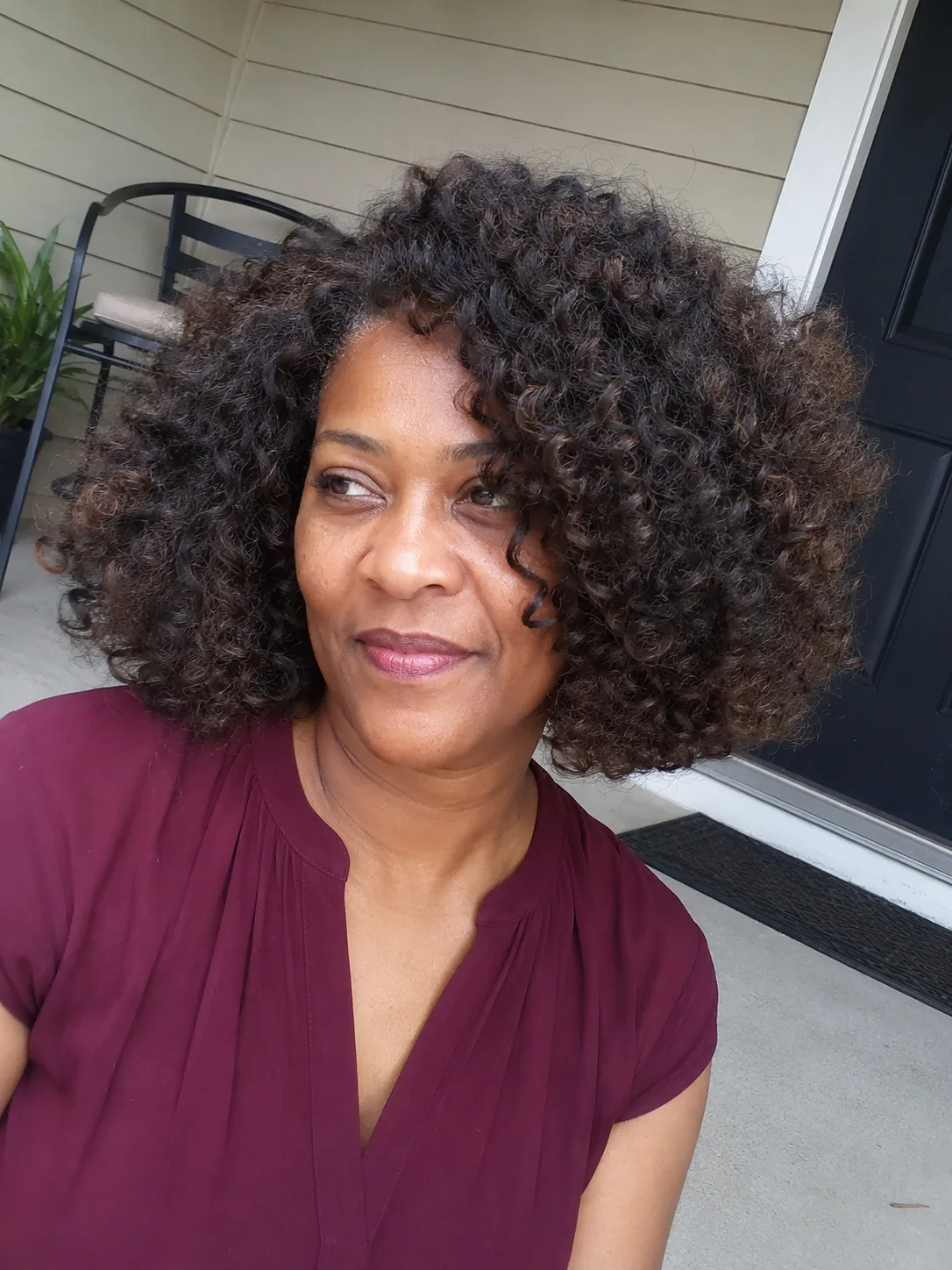 A casual indoor portrait of a woman with long, romantic auburn-brown curls parted down the middle, showcasing soft, defined tendrils cascading over her shoulders
