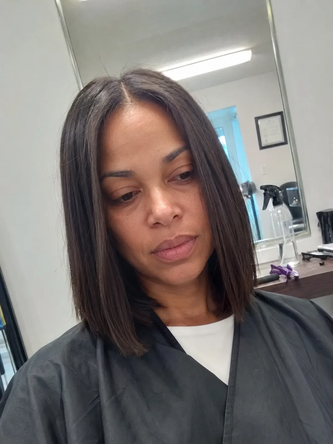 A close-up portrait of a woman inside a salon, displaying a sleek, straight, center-parted dark brown lob (long bob) haircut that rests just above her shoulders