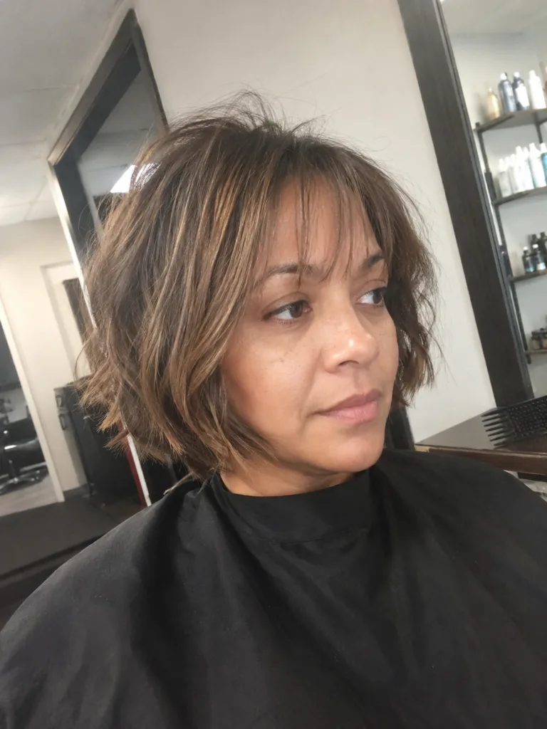 A close-up portrait of a woman with a messy, textured brown pixie cut featuring soft, layered bangs framing her forehead naturally.