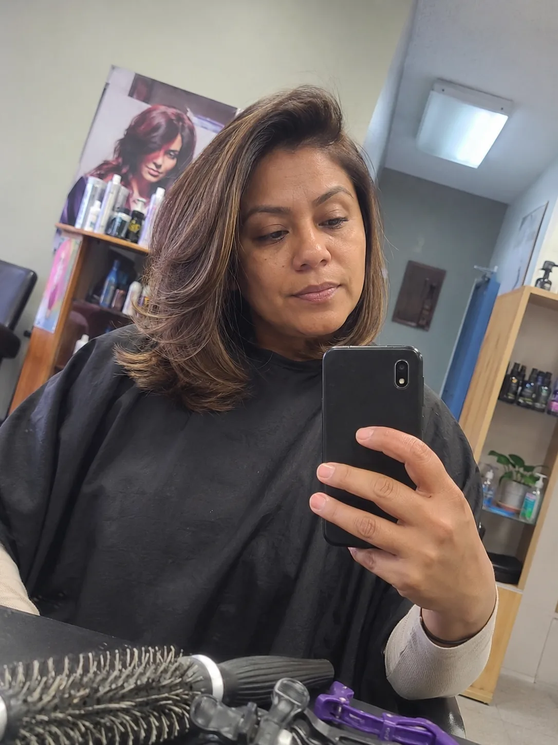 A mirror selfie of a woman in a salon cape, showcasing a voluminous, side-swept medium-length blowout with flipped-out layers and warm honey-brown highlights