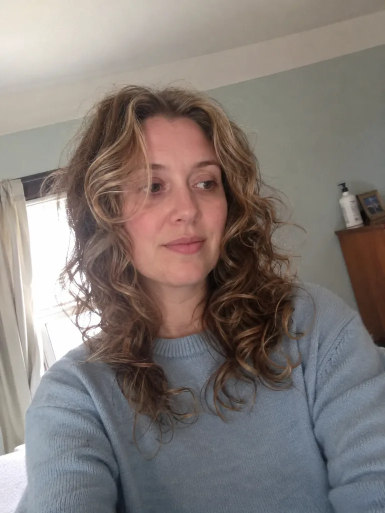 A portrait of a woman looking directly forward, showcasing voluminous, layered dark curls swept over one shoulder with subtle golden-brown highlights.