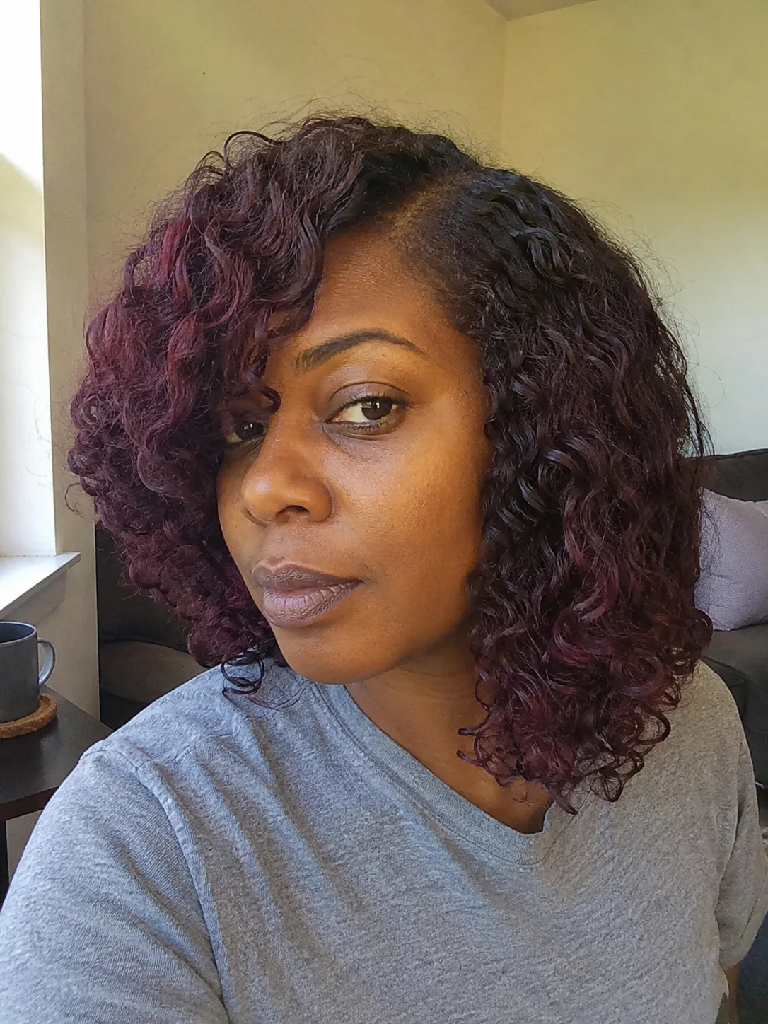 A portrait of a woman looking toward the camera, highlighting textured curly hair dyed a deep plum and dark burgundy hue