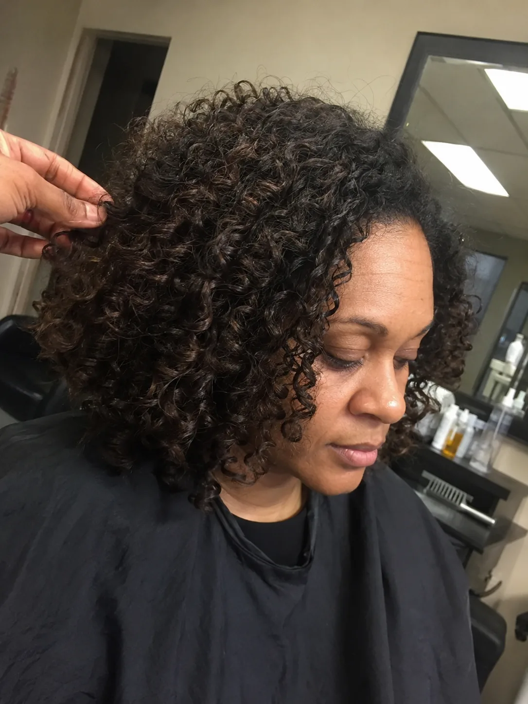 A portrait of a woman sitting in a salon chair, featuring shoulder-length, loose dark wavy hair styled in a layered shag cut with effortless volume