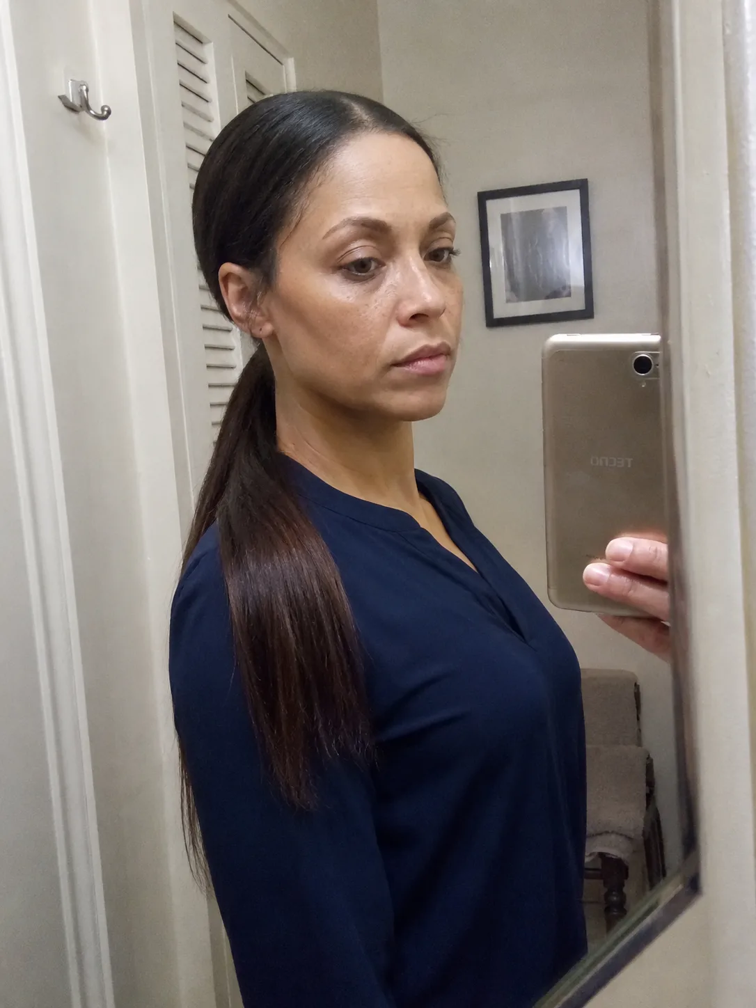 A profile view mirror selfie of a woman wearing a blue blouse, showcasing long, sleek, and smooth dark brown hair pulled back into a low ponytail