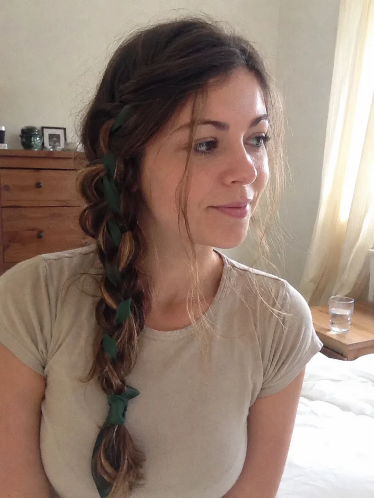 A side profile of a woman with long, thick brown hair styled in a loose Celtic braid adorned with small white and pink wildflowers.