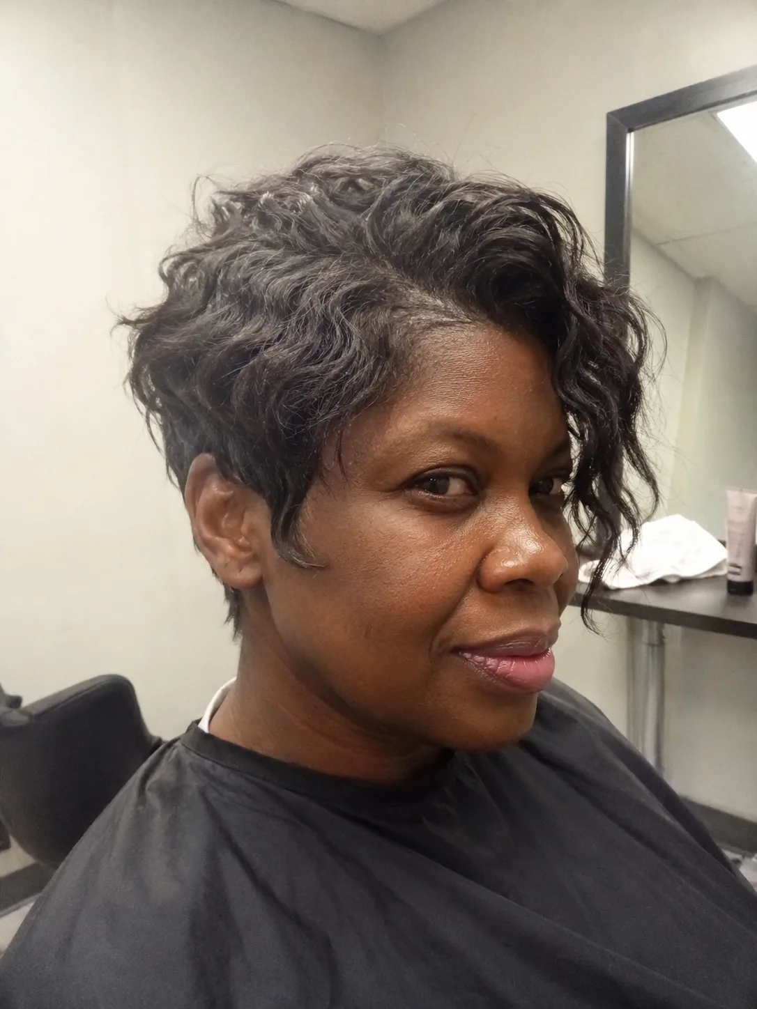 A side-profile shot of a woman in an office setting showing her long, dark highlighted hair gathered into a thick, textured low ponytail with loose waves