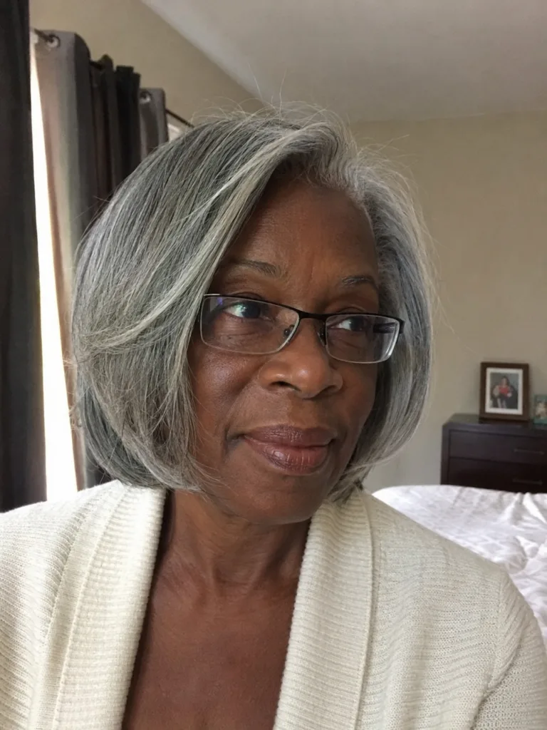 A woman with glasses sitting on a sofa, displaying long, layered flowing grey hair with natural dark undertones.