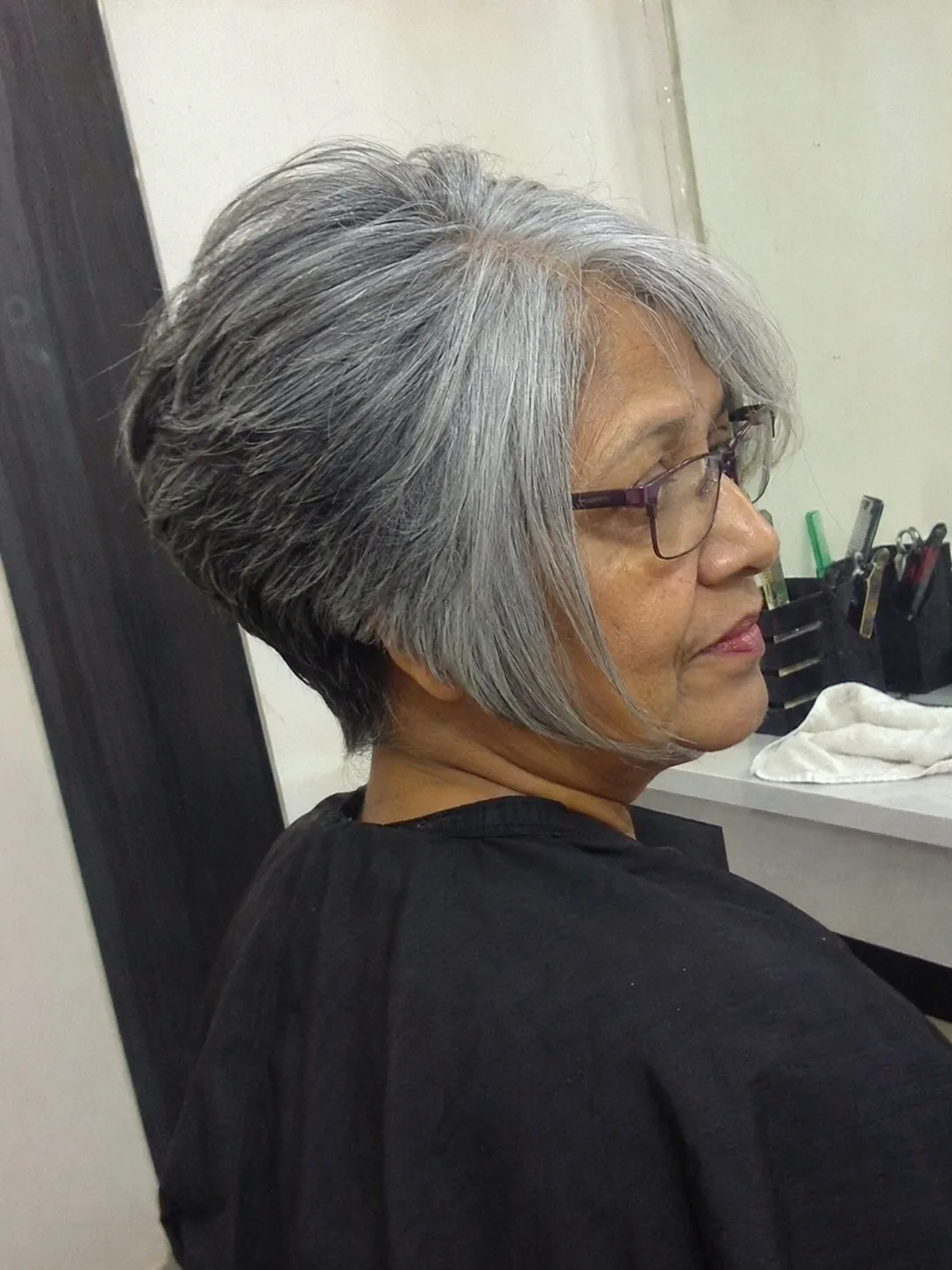 Side profile view of a woman in a salon setup, showing a structured, angled inverted grey bob haircut paired with glasses