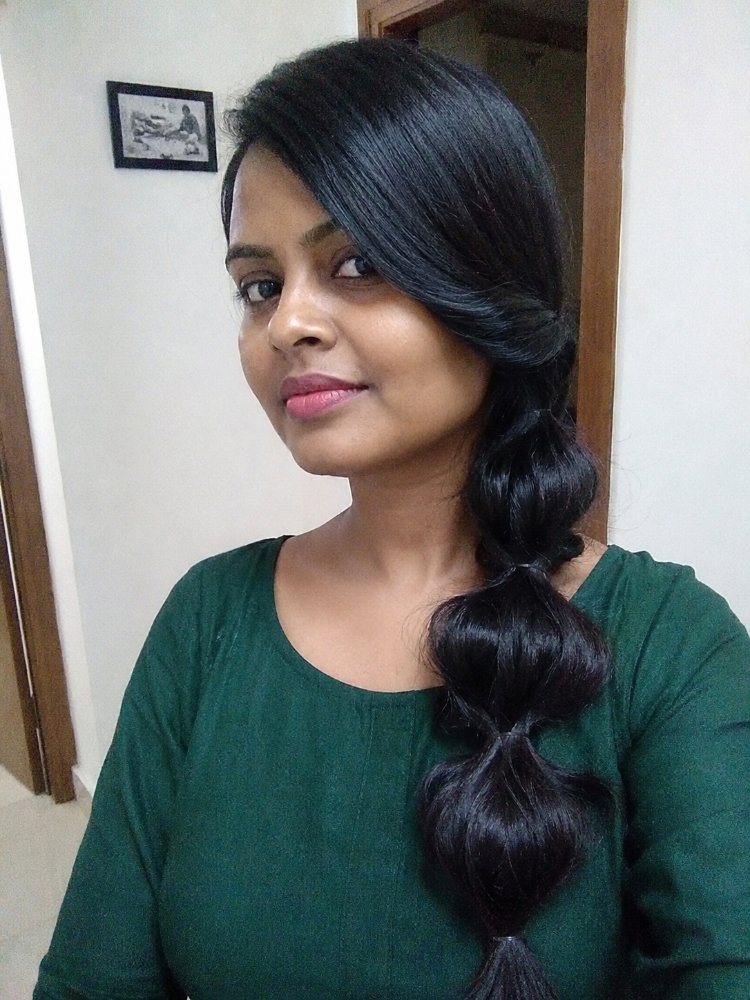The hair is dark brown-black in color, long in length, and smooth in texture. It is styled in a loose, bubble-like braid that falls down the back, with face-framing strands swept to the side, and appears to have a subtle sheen, but