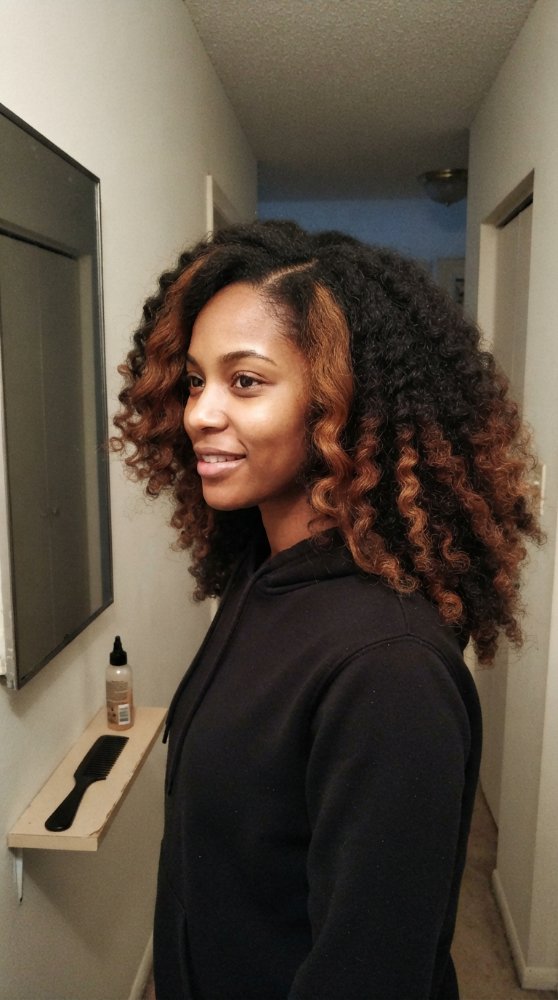 The hair is dark brown with caramel-colored highlights, featuring loose, spiral curls that fall just below the shoulders