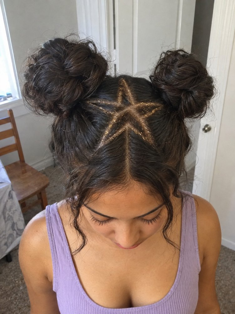 dark brown color styled two The hair is dark brown in color, styled into two space buns on either side of the head. A star design is created with glittery hair product on the crown, starting from the center part and extending outwards in a zig-zag pattern, adding dimension and visual interest to the overall look. The hair appears to be of medium length and has a slightly textured appearance
