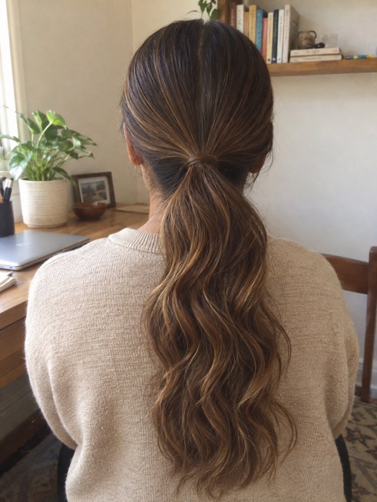 dark brown color subtle highlights The hair is a dark brown color with subtle highlights of lighter brown and blonde, creating dimension and depth. It appears to be long, reaching down to the middle of the back, with a wavy texture and loose curls that are more pronounced towards the ends. The hair is styled in a low ponytail with a center part