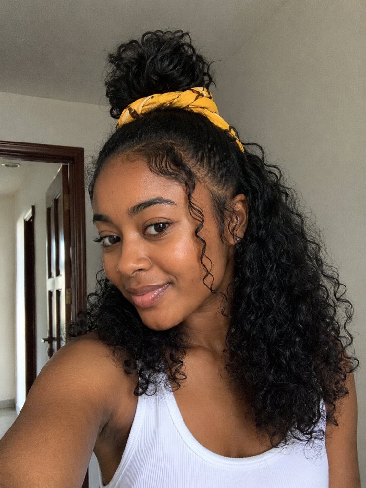 dark brown curly texture appears The hair is dark brown with a curly texture and appears to be long, reaching down to the mid-back. The top section is pulled up into a high bun and secured with a yellow bandana, while the lower section cascades down in loose, bouncy curls with a few face-framing tendrils