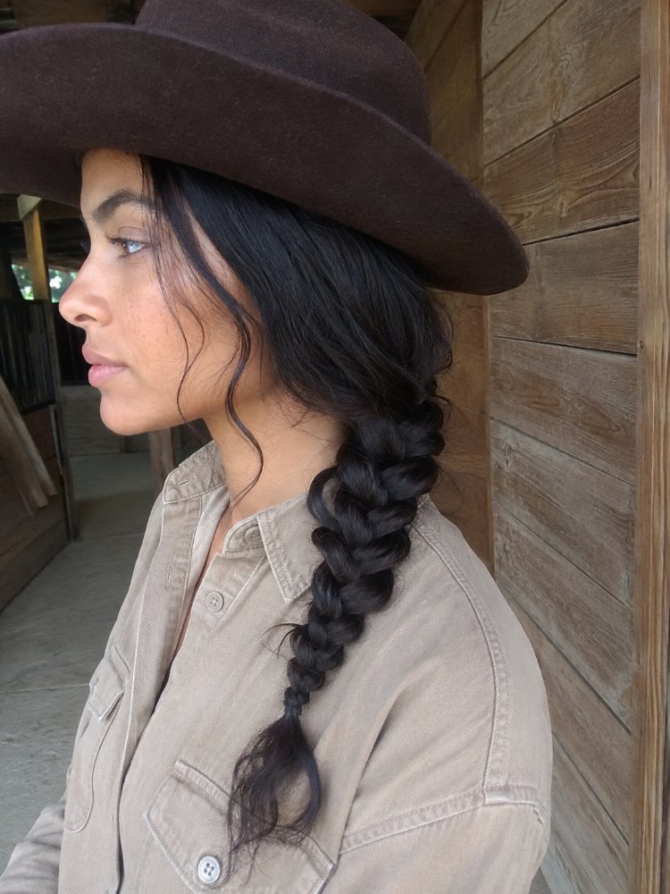 dark brown falls long thick The hair is dark brown and falls in a long, thick braid down the back. The braid appears to be a classic French braid, with a smooth texture and visible tangles or knots, and some loose strands frame the face. The hair color is a uniform dark brown with subtle highlights, and has a sleek, shiny appearance