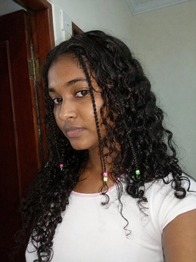 dark brown falls medium length The hair is dark brown and falls to a medium length. It has a curly texture and is styled in small braids with beads at the ends, adding dimension and visual interest. The braids frame the face and have a relaxed, wavy pattern that adds movement and texture to the overall hairstyle