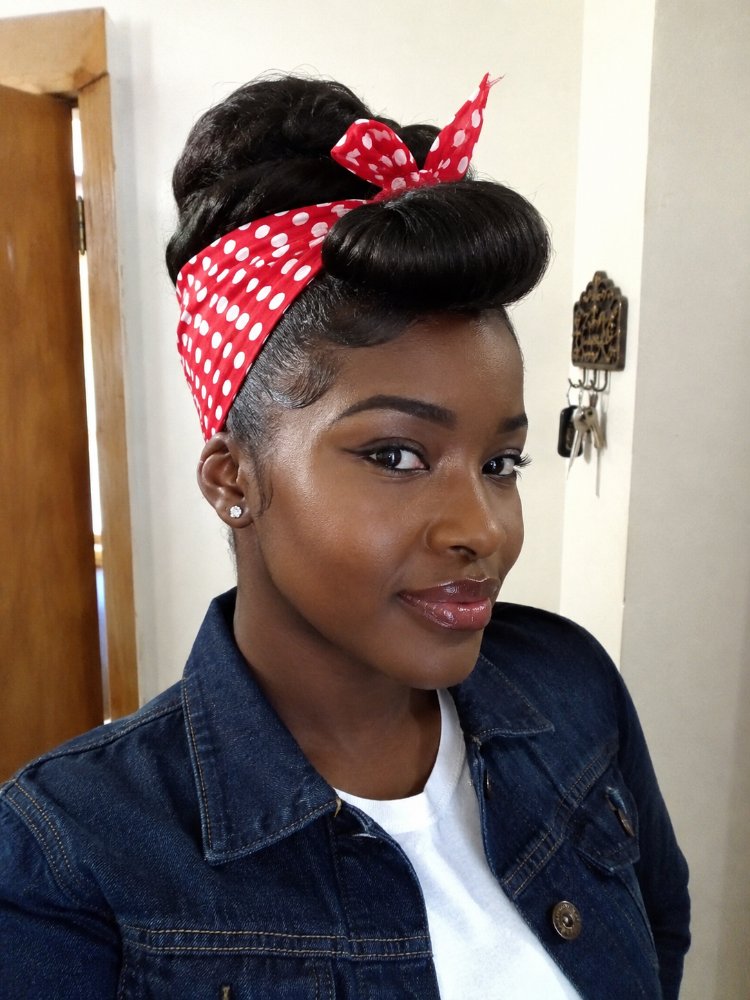 The hair is dark brown and styled in an updo, with a sleek, smooth texture. The hair is pulled back into a high bun, with a few strands framing the face, and secured with a red bandana featuring white polka dots, which adds a pop of color and texture. The overall style is polished and sleek, with visible layered styling, and appears to have been achieved with the use of styling products to achieve a glossy finish