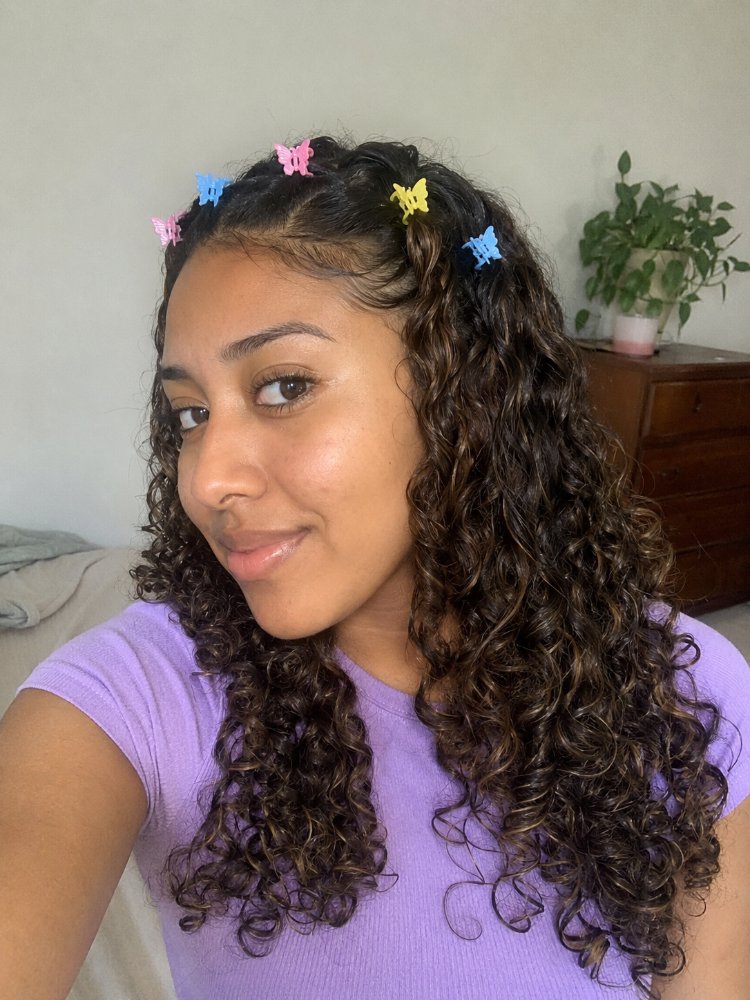 dark brown subtle highlights featuring The hair is dark brown with subtle highlights, featuring tight, loose curls that fall to a medium length. The top section of hair is partially pulled back and adorned with small, colorful butterfly clips, while the rest of the hair cascades down, showcasing its natural texture and dimension