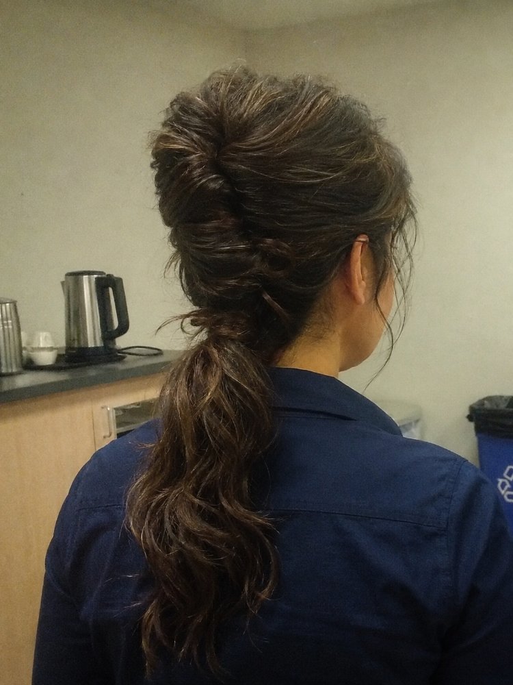 The hair is dark brown with subtle highlights of lighter brown, and its length falls down the back in a low ponytail that reaches the middle of the back.