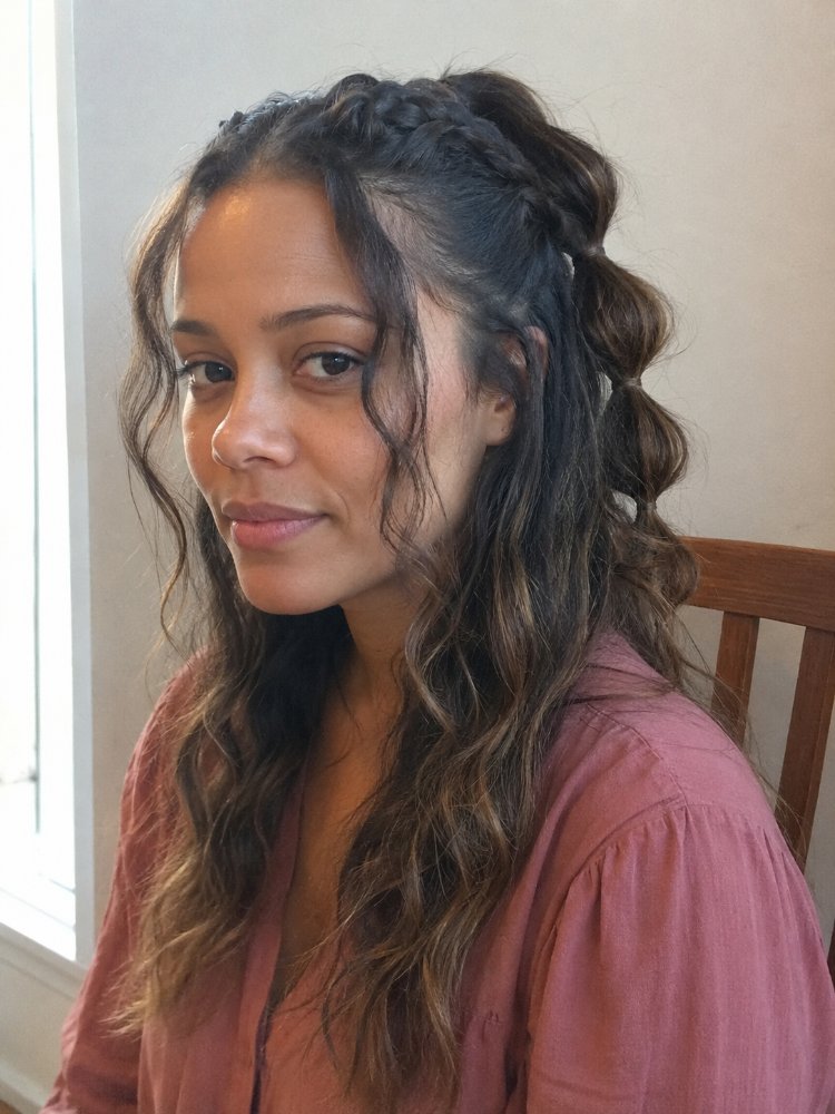 The hair is dark brown with subtle highlights of lighter brown and blonde, adding dimension and texture. It falls in loose, wavy curls down to a medium length, with a braided crown that adds a touch of elegance. The hair appears to have a natural, effortless look with a few stray strands framing the face