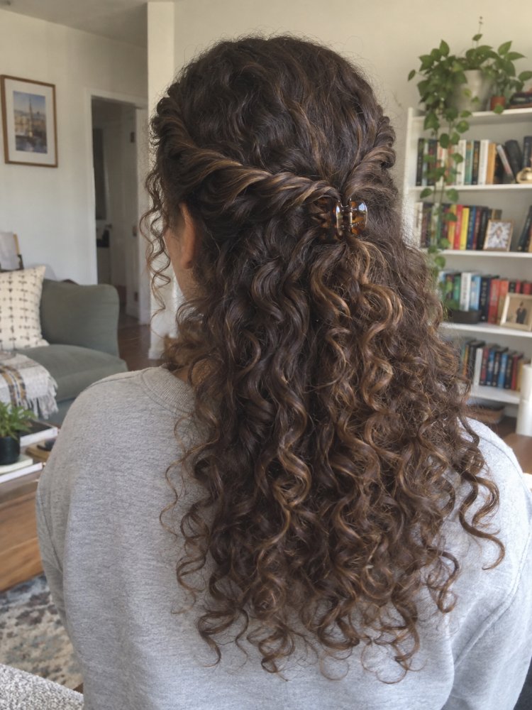 dark brown subtle highlights lighter The hair is dark brown with subtle highlights of lighter brown, creating dimension and depth. It appears to be long, reaching down to the middle of the back, with a curly texture that is loose and flowing, and a half-up style with a braided section framing the crown. The curls are tight and bouncy, with natural color transition