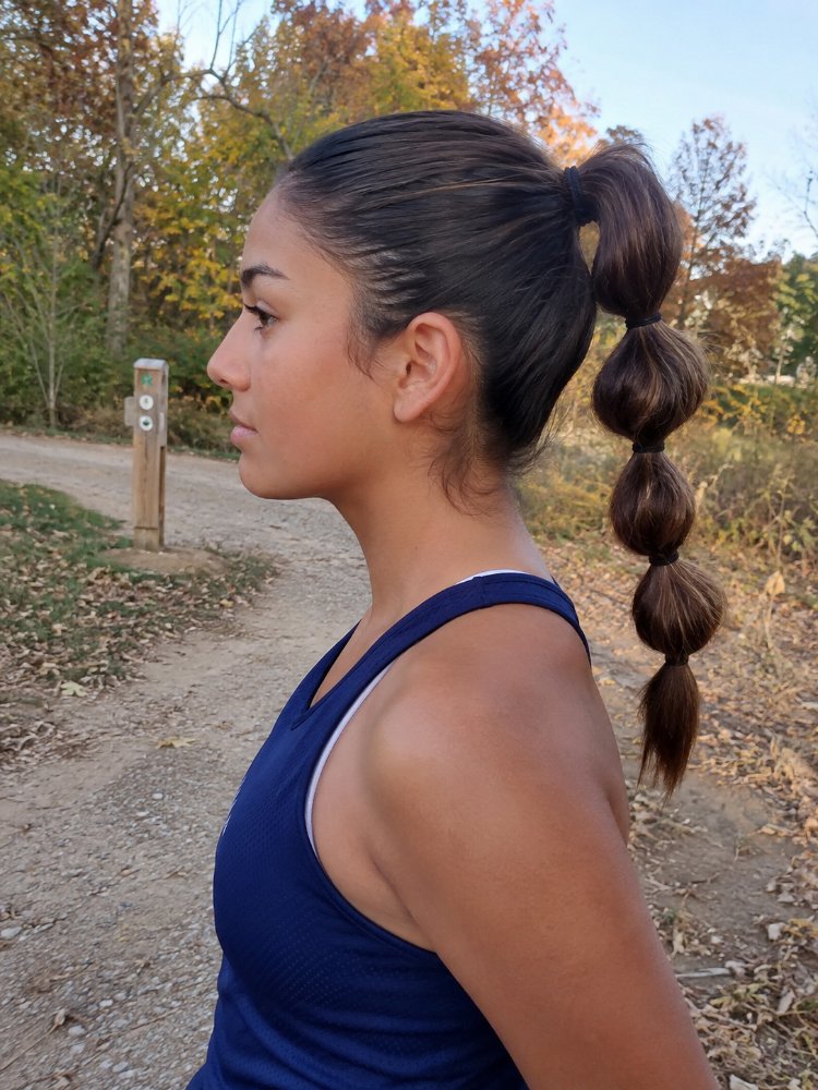dark brown subtle hints lighter The hair is dark brown with subtle hints of lighter brown, styled in a unique bubble ponytail with four distinct sections. The length appears to be past the shoulders, and the texture seems smooth and sleek, with visible curls or braids. The cut style is not clearly visible, but it appears to be a simple, straight cut with layers framing the face