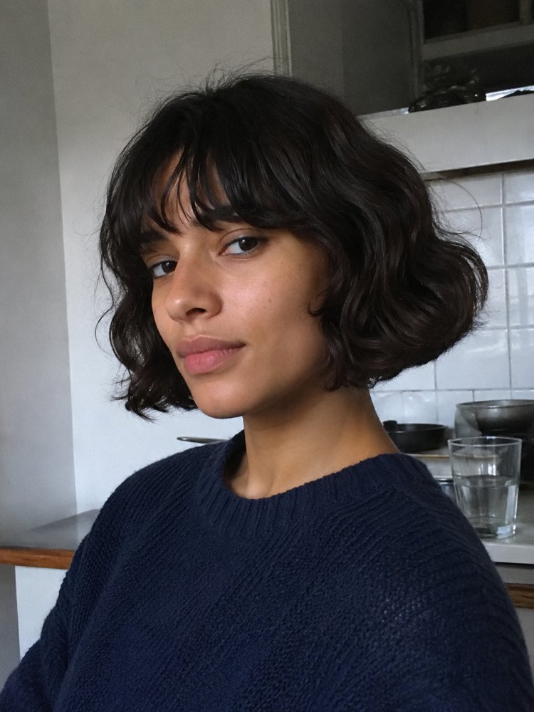 dark brown subtle natural looking texture The hair is dark brown with a subtle, natural-looking texture. It is cut in a short, chin-length bob with choppy layers and blunt bangs that frame the face, adding dimension to the overall look. The hair appears to be free of curls, braids