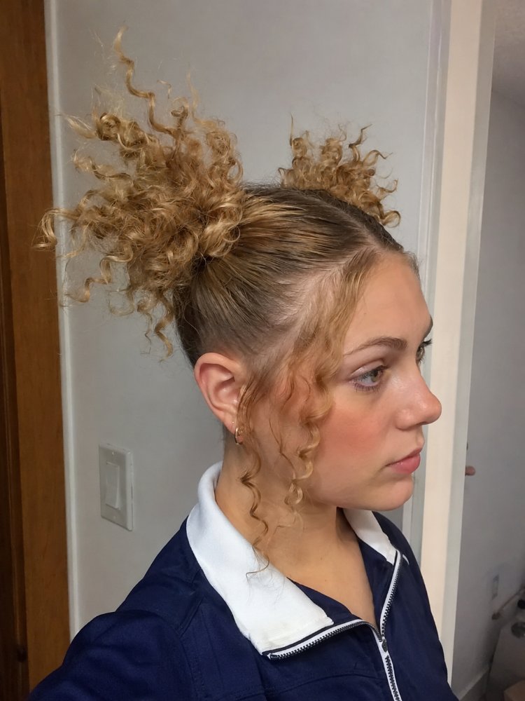 light blonde color darker roots The hair is a light blonde color with darker roots, styled into a messy, curly high ponytail. The curls are loose and bouncy, with some tendrils framing the face, and the texture appears to be slightly frizzy and piecey. The hair length is medium to long, with layers that add to the volume and movement of the curls