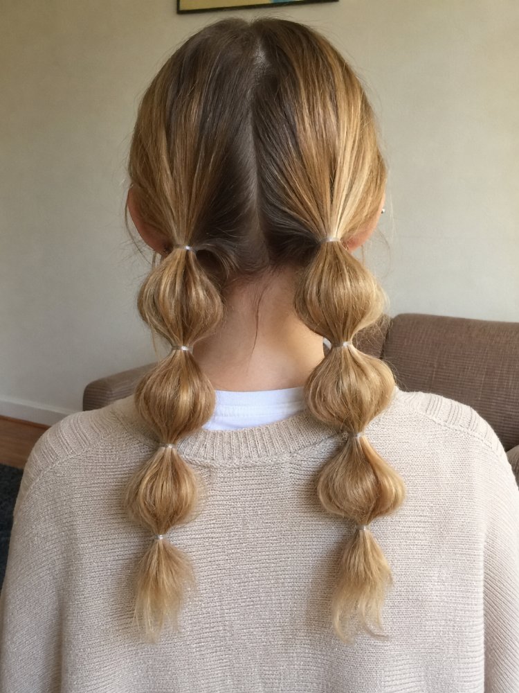 The hair is a light blonde color with darker roots, and appears to be long, reaching down to the middle of the back. The texture seems smooth and silky, with a subtle sheen; it is styled in two bubble braids with small clear elastic bands separating the sections, and has a face-framing effect around the neckline