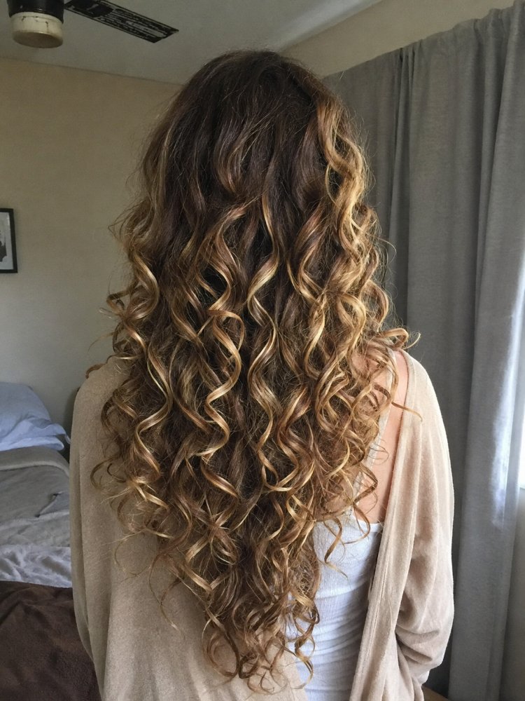 long extending down lower back The hair is long, extending down to the lower back, with a rich, dark brown base color that transitions into a multi-dimensional blonde ombre effect. The locks feature loose, spiral curls that cascade down, adding movement and texture, with subtle highlights and lowlights that create a natural, sun-kissed look, likely achieved through balayage