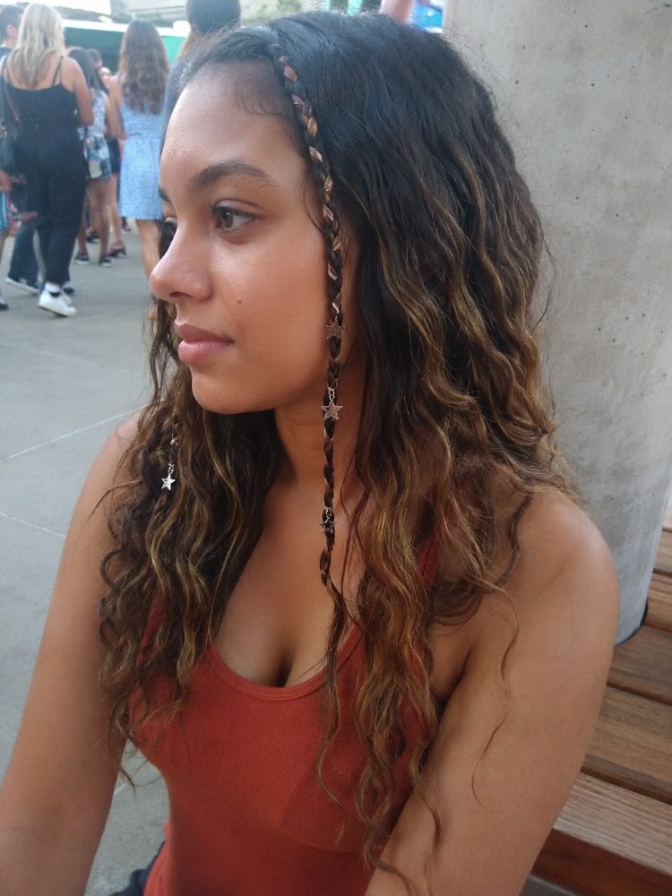 long wavy dark brown subtle The hair is long, wavy, and dark brown with subtle highlights of lighter brown and blonde tones, giving it dimension and depth. Two small braids frame the face, each adorned with tiny star-shaped charms, adding a decorative touch. The hair appears to have a natural, effortless texture with loose curls and subtle layering