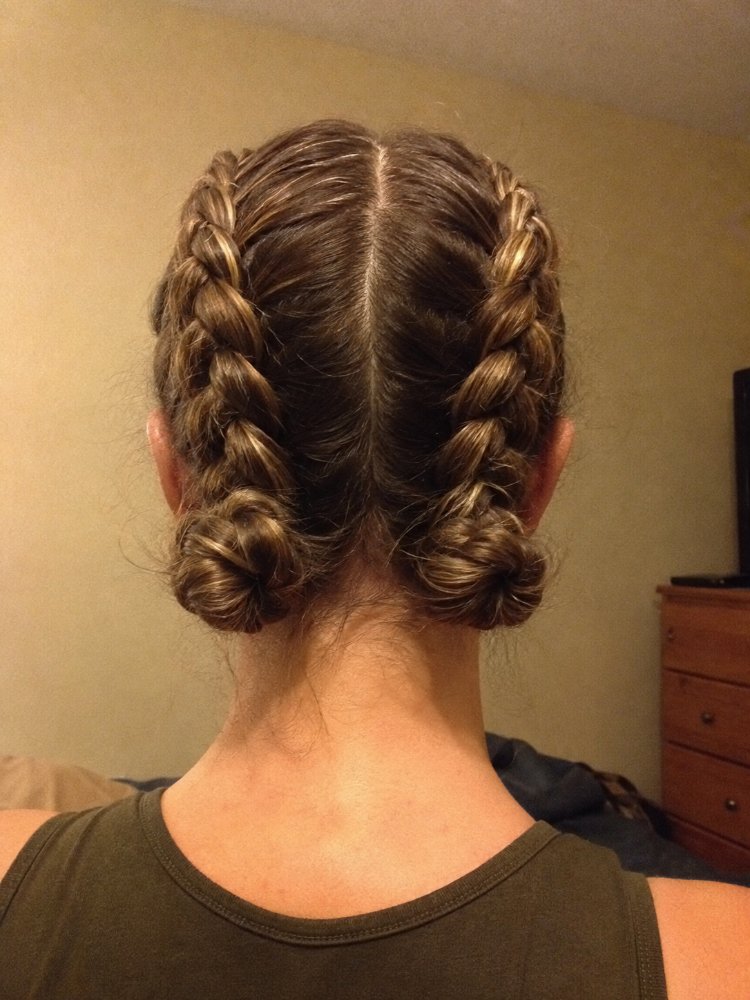 medium dark brown color subtle The hair is a medium to dark brown color with subtle highlights of lighter brown and blonde tones. It is styled in two French braids that are coiled into buns at the nape of the neck, with a center parting visible at the roots. The hair appears to be of medium length