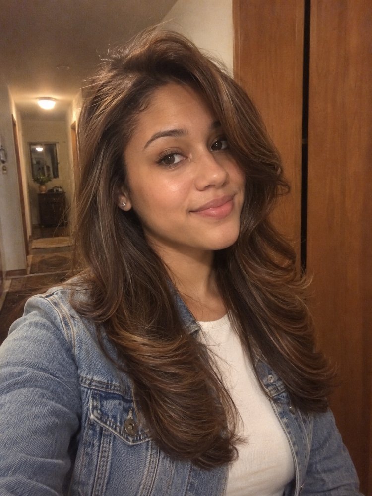 The hair is a rich, dark brown color with subtle highlights that add dimension and texture. It falls in loose, flowing waves that reach mid-length, framing the face with layers that enhance movement and flow. The hair appears to have been styled with a gloss or toner to achieve a smooth, healthy-looking finish
