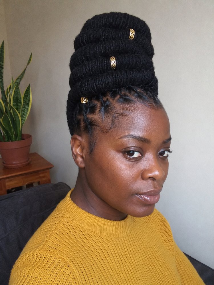 The hair is styled in a tall, braided updo with a dark brown or black color. The braids are thick and textured, with a few gold beads woven throughout, and appear to be tightly coiled, giving a dimensional look. The style features face-framing braids around the sides and back of the head, which are swept up into the tall bun, with visible layers, curls or regrowth, and dimensional color